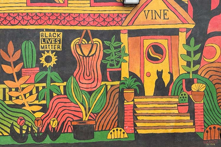 Solstace is a mural painted by Travis Grabill that depicts a black cat sitting on a porch of a typical house found in the Vine Street Neighborhood. The scene uses oranges, reds, yellows, black and green to celebrate the arrival of summer.